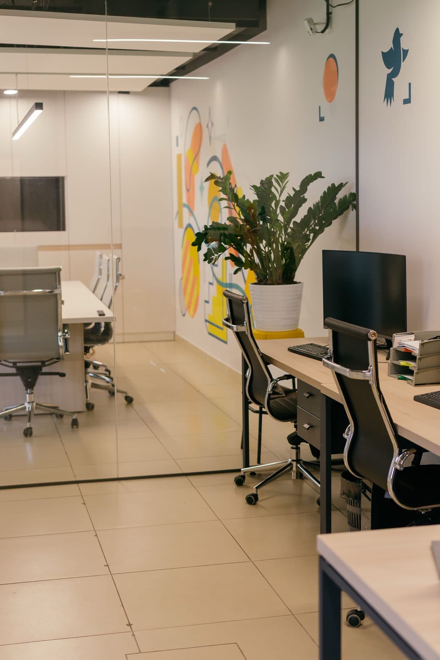 An empty open-plan office with desks, chairs, a potted plant, and a colorful mural
